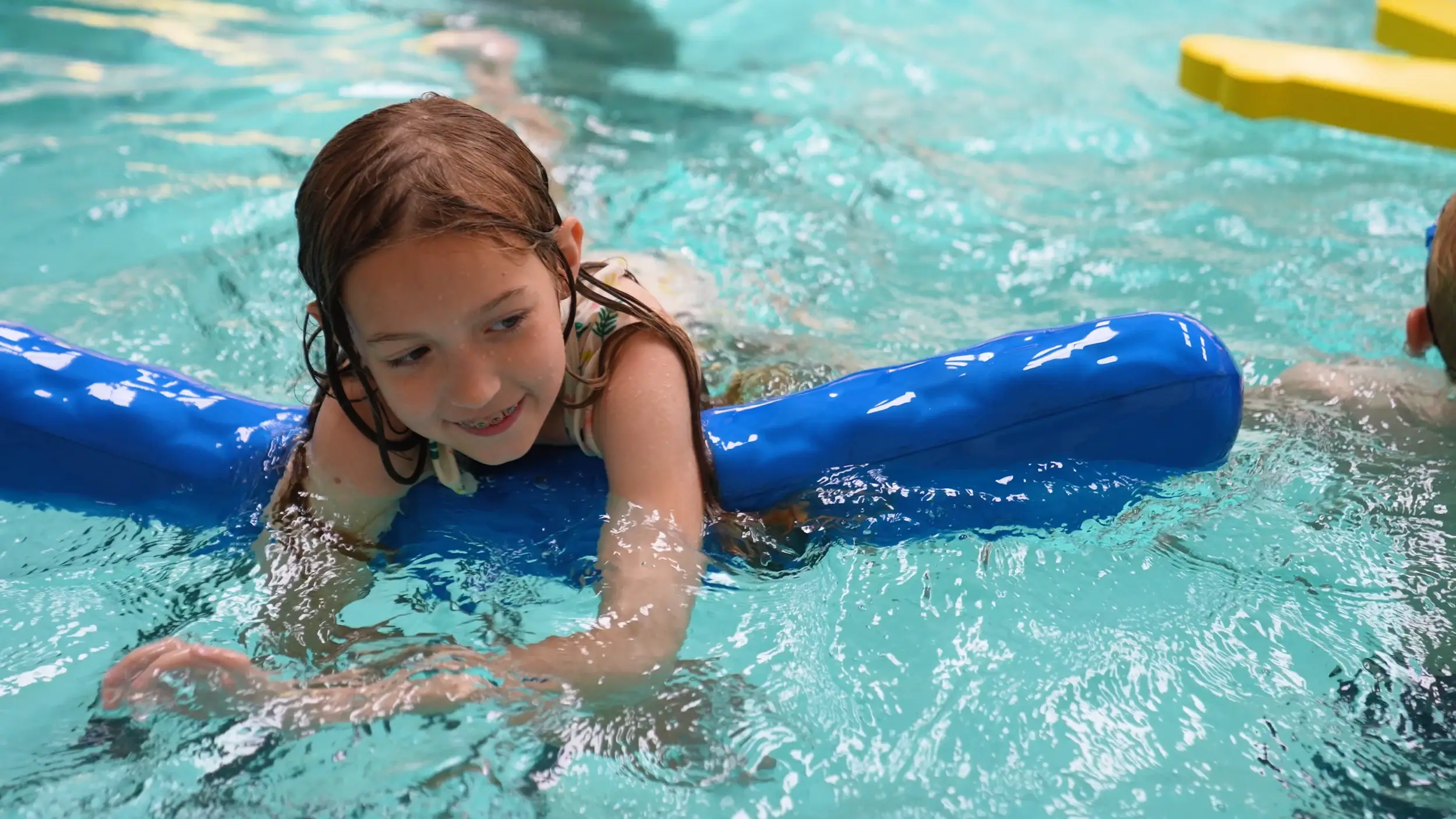 A young swimmer practices over a blue float with an instructor nearby
